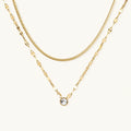 Layered gold necklace with herringbone and link chains, small bezel-set crystal pendant on cream background