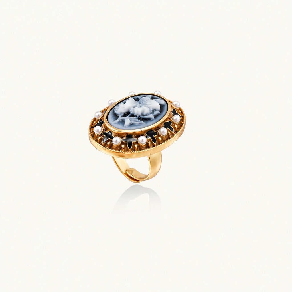Gold-plated ring with oval blue floral cameo, pearl accents, and black enamel detailing on white background.