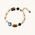 Gold chain bracelet with tiger's eye, obsidian, pearl, and iridescent abalone stones on cream background.