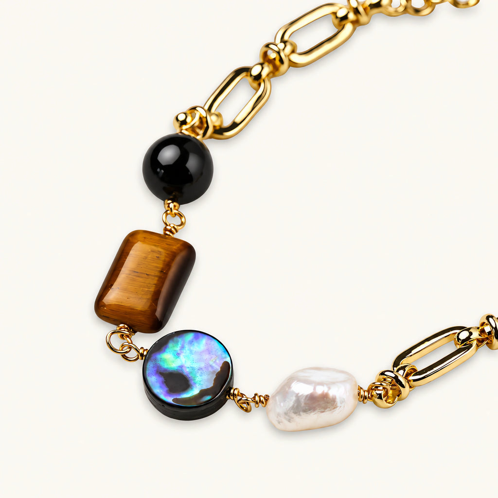 Gold chain bracelet with tiger's eye, obsidian, pearl, and iridescent abalone stones on cream background.